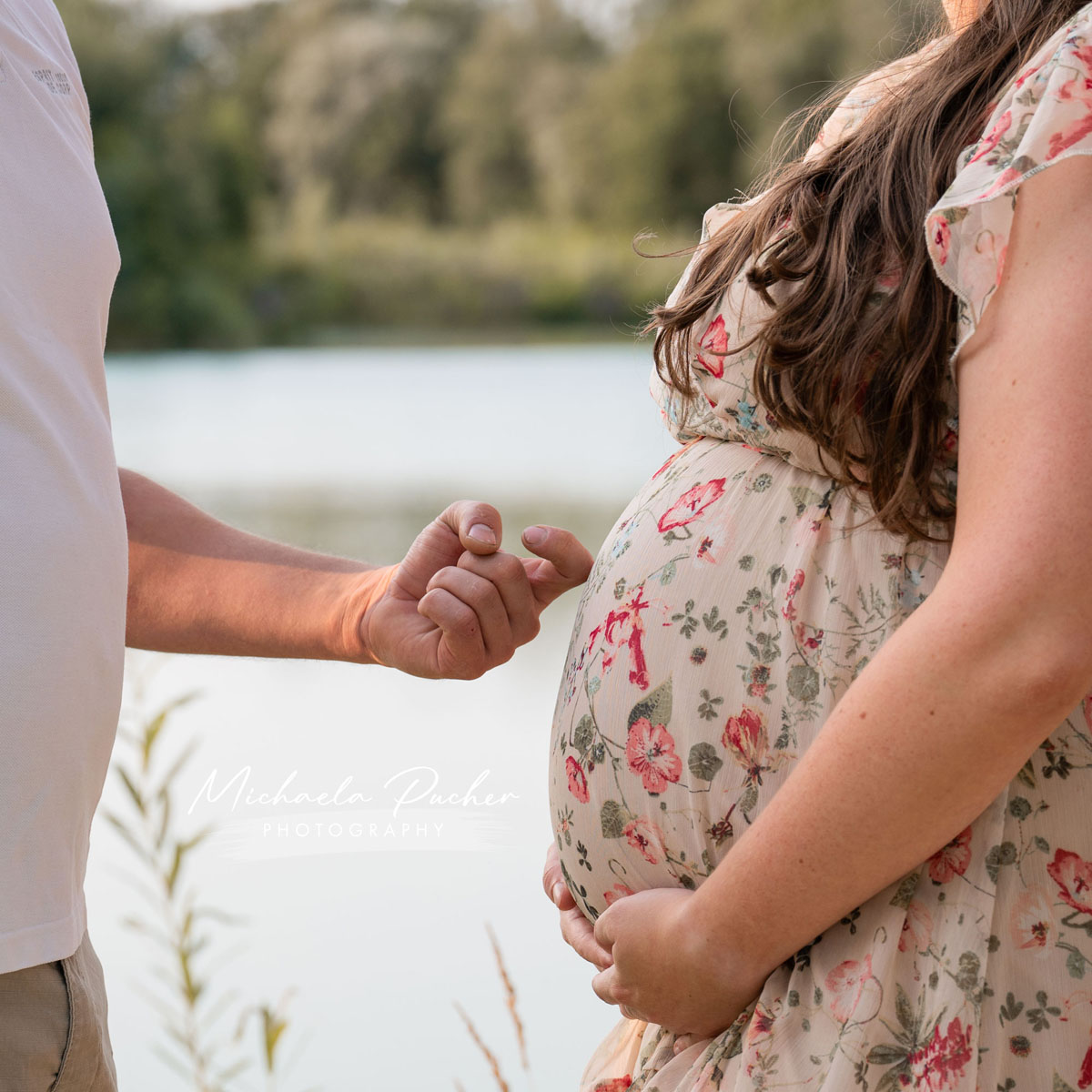 Nahaufnahme eines Babybauchs im geblümten Kleid, während eine Hand des Partners liebevoll den Bauch berührt und die Sonne eine warme Stimmung am See erzeugt.